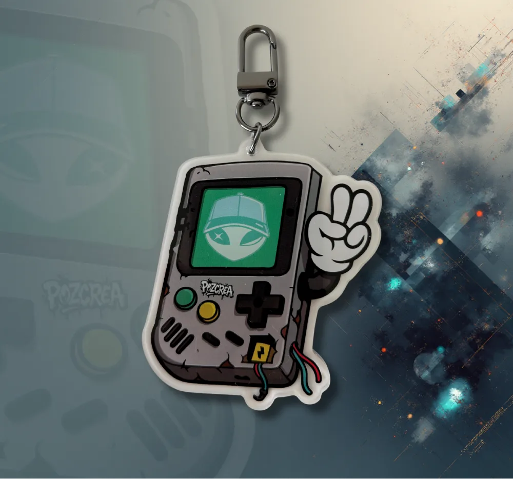 Porte-clé GameBoy Alien vu de face, posé à plat. L’écran affiche toujours le visage de l’alien, avec une main levée en signe de victoire. Les boutons sont visibles, et le crochet noir est positionné en haut.