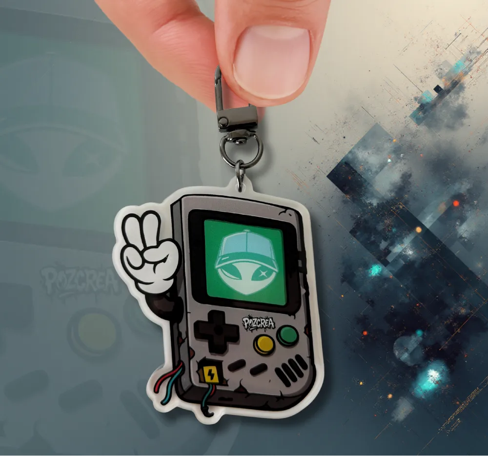 Porte-clé en forme de GameBoy tenu entre deux doigts. Sur l’écran vert se trouve le visage de l’Alien POZCREA, et la console affiche un bras qui fait le signe "peace". Les boutons sont visibles, le fond est flou et sombre avec des reflets bleus.